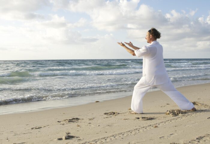 Bilden visar en person på en strand som utövar tai chi eller yoga.